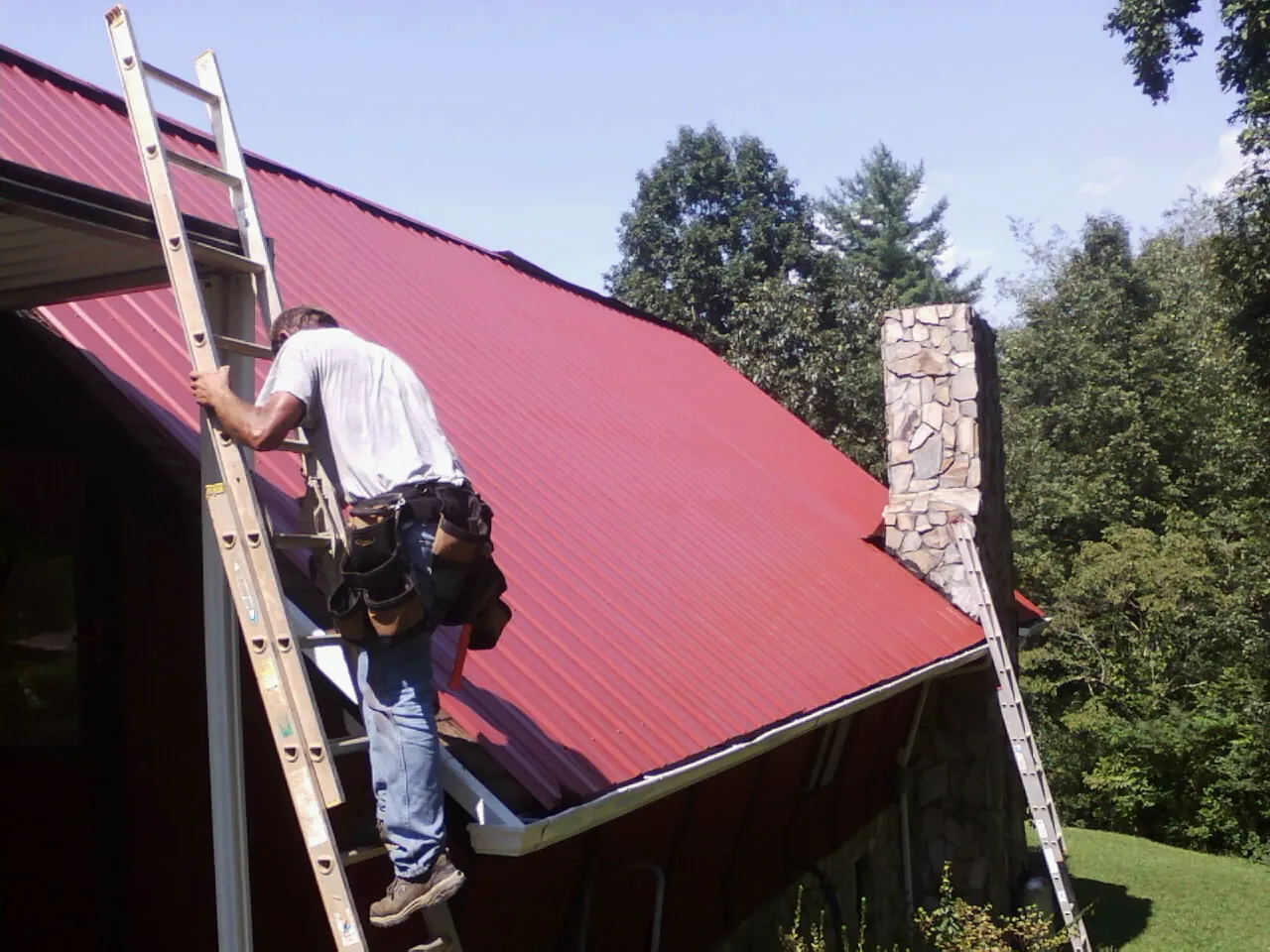 Roofer on a ladder beside a red metal roof and stone chimney — Soffit & Fascia Repair by Roofing Brothers in Smock
